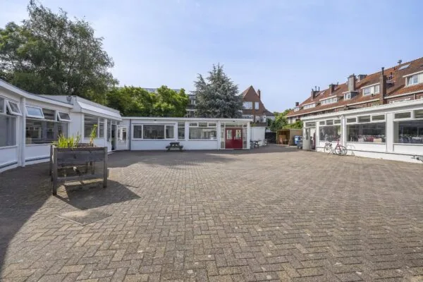 Binnenplaats met bestrating en lage witte school- of kantoorgebouwen aan de Jacques Perkstraat, omringd door woningen en groen.